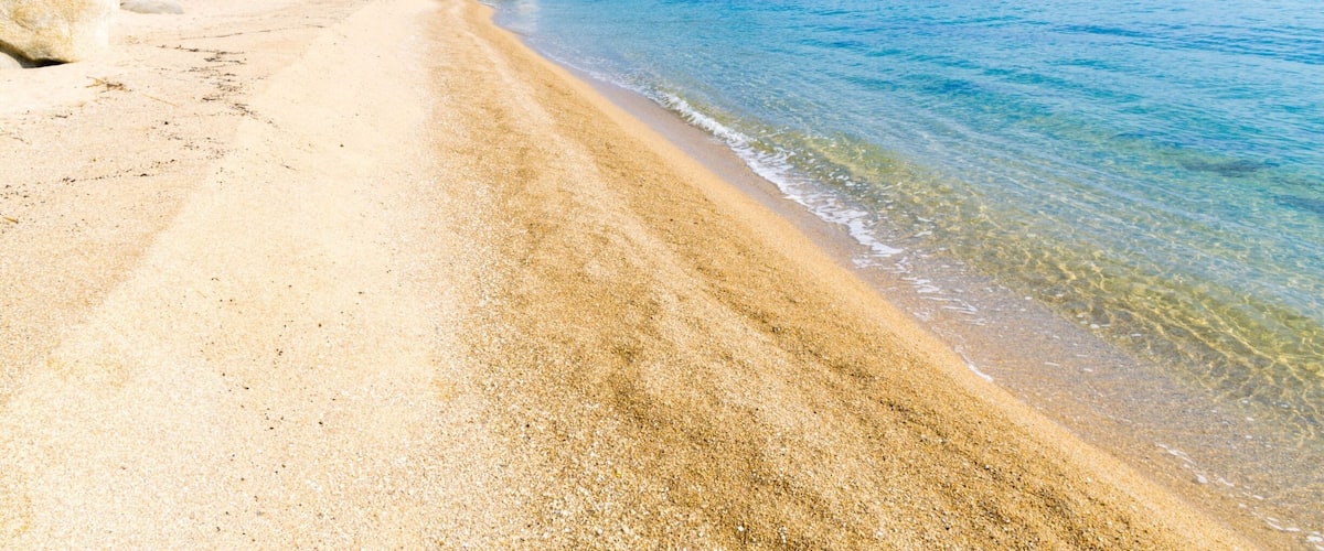 A sandy beach with clear water, rocky cliffs, and green hills in the background.