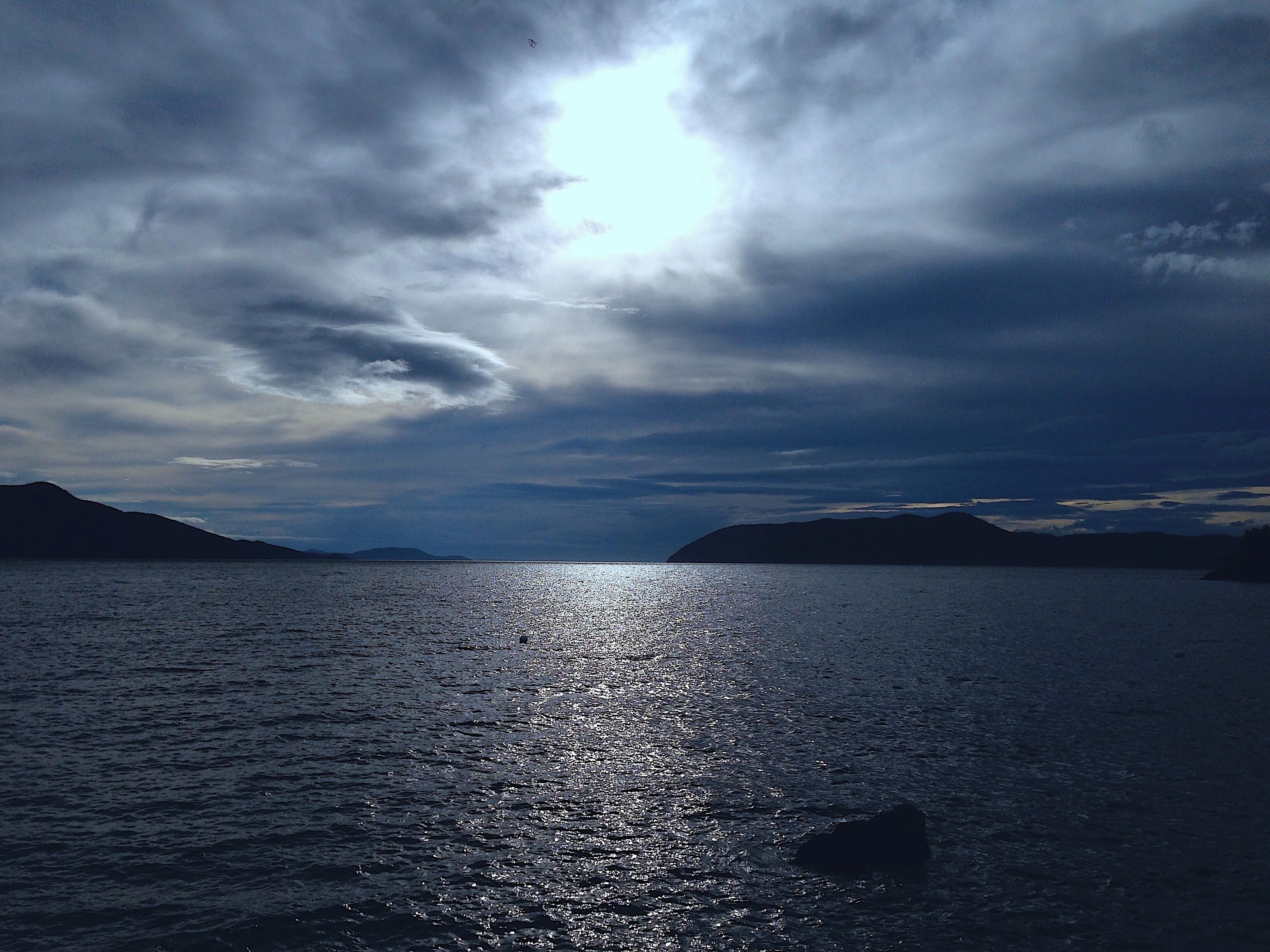 A calm sea with sunlight reflecting off the water, surrounded by mountains and a cloudy sky.