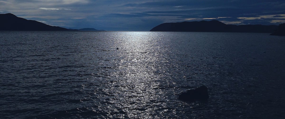 A calm sea with sunlight reflecting off the water, surrounded by mountains and a cloudy sky.