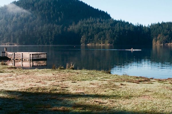 Ein ruhiger See mit einem hölzernen Steg und einem bewaldeten Hügel im Hintergrund.
