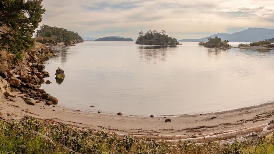 A calm lake with a sandy beach, surrounded by rocky cliffs and autumn-colored trees.