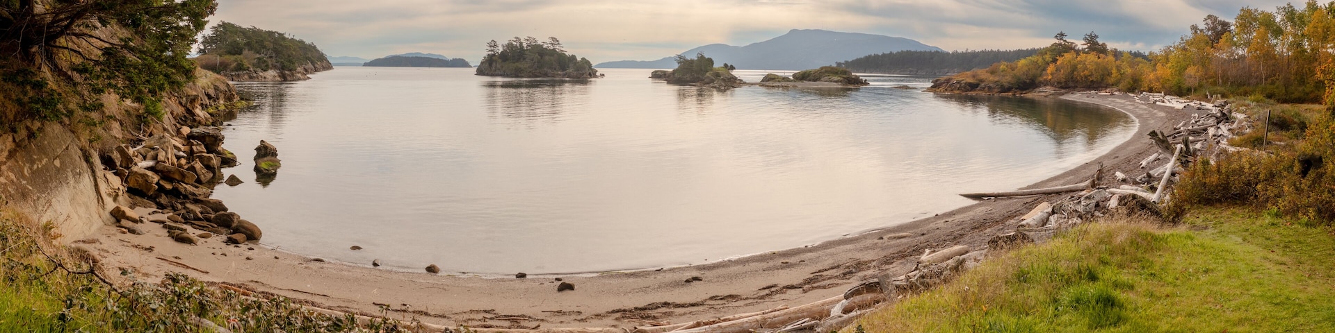 Ein ruhiger See mit einem Sandstrand, umgeben von felsigen Klippen und Bäumen in Herbstfarben.