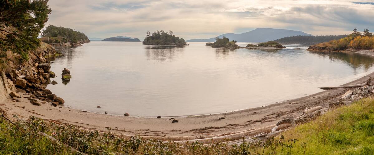 A calm lake with a sandy beach, surrounded by rocky cliffs and autumn-colored trees.