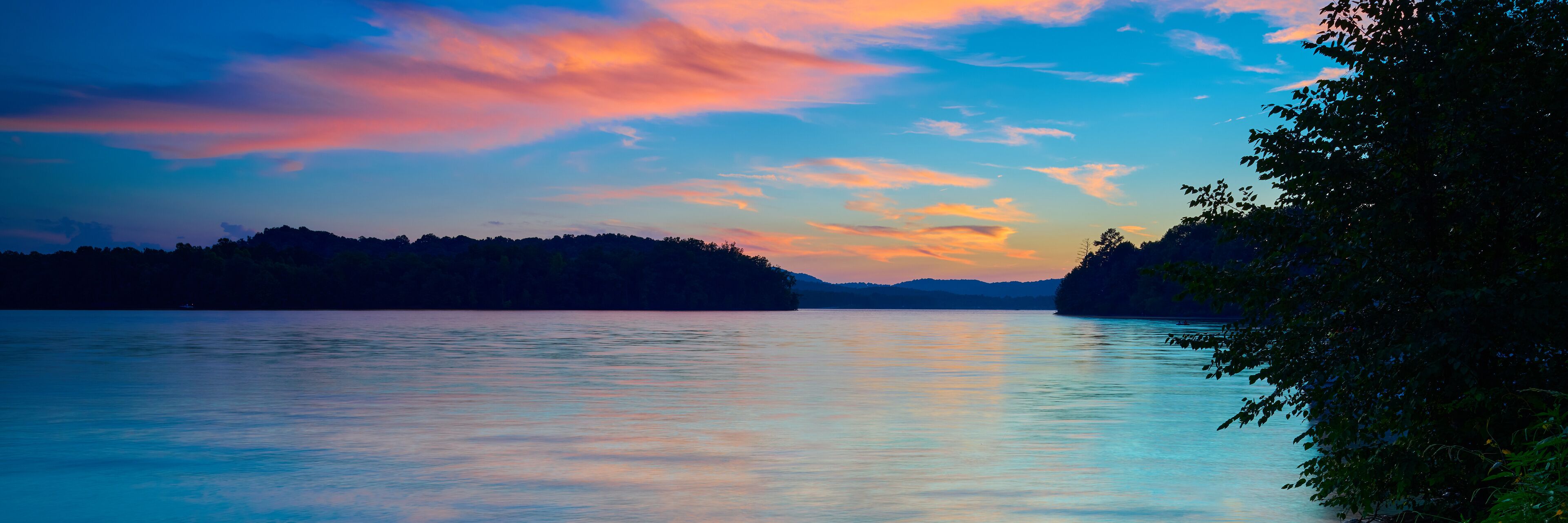 A calm lake at sunset with a forested shoreline and a colorful sky.