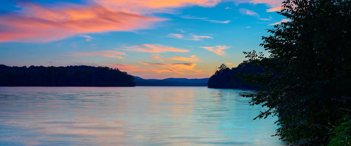 A calm lake at sunset with a forested shoreline and a colorful sky.