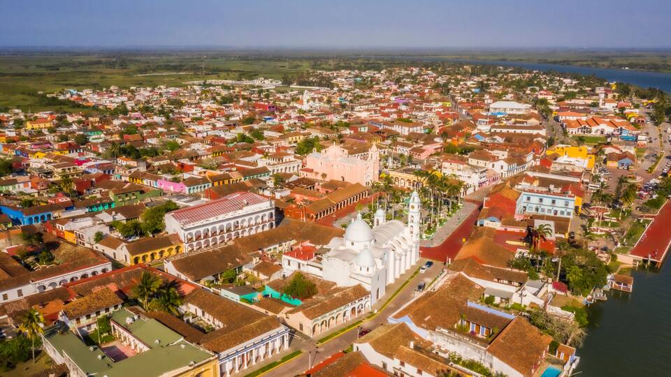 An aerial view of a coastal town with colorful buildings, a prominent white church with a bell tower, and a body of water to the right.