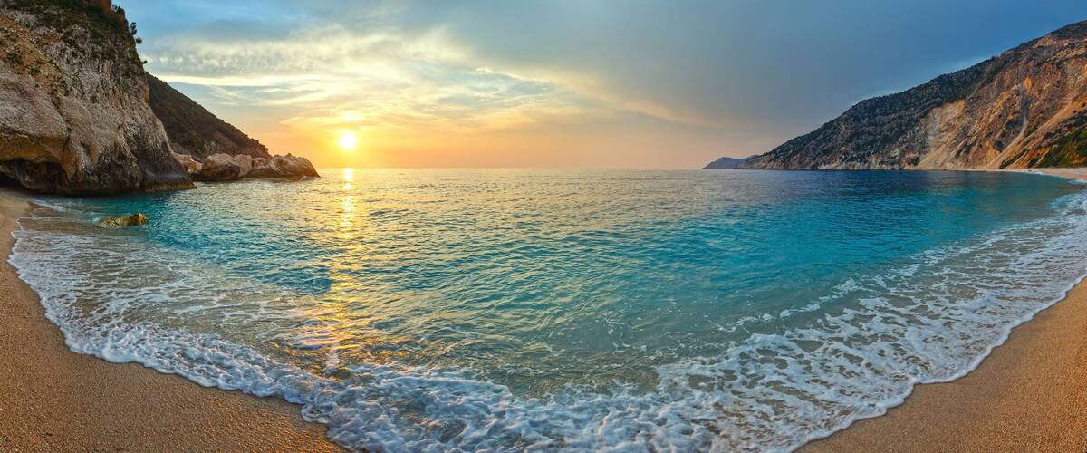 A beach with clear turquoise water, a sandy shore, and rocky cliffs under a sunset sky.