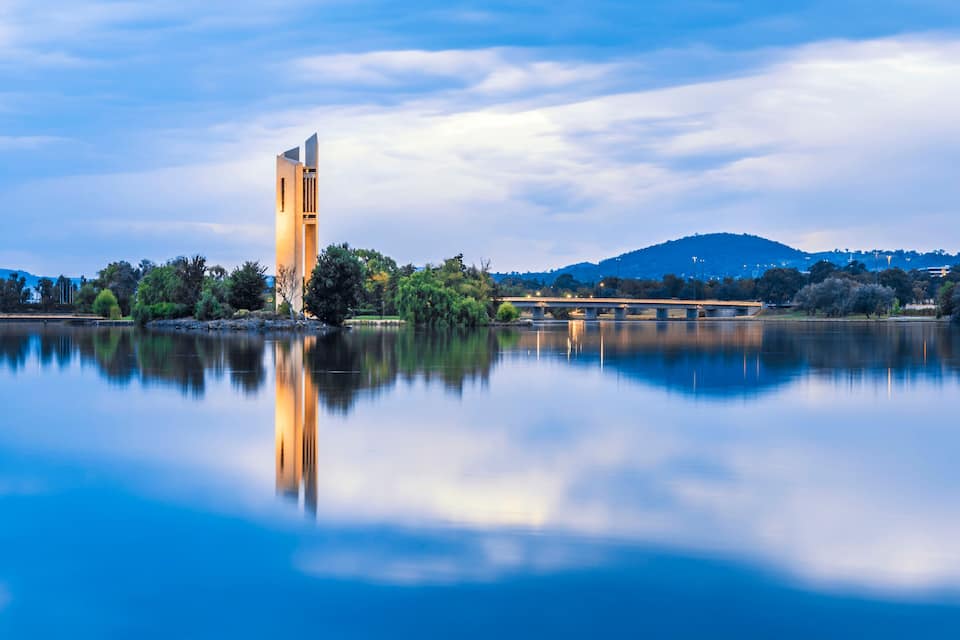 A calm lake with a tall tower reflecting on the water, surrounded by trees and a bridge in the background.