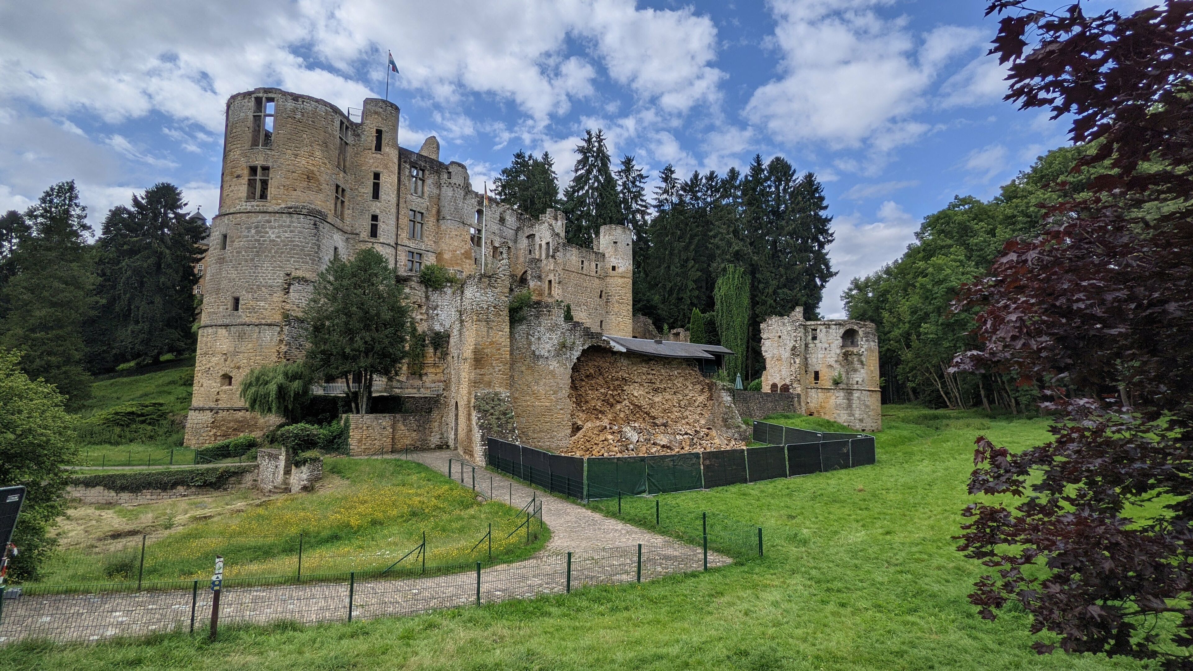 Un castillo histórico con muros y torres de piedra, rodeado de árboles y un espacio cubierto de césped.