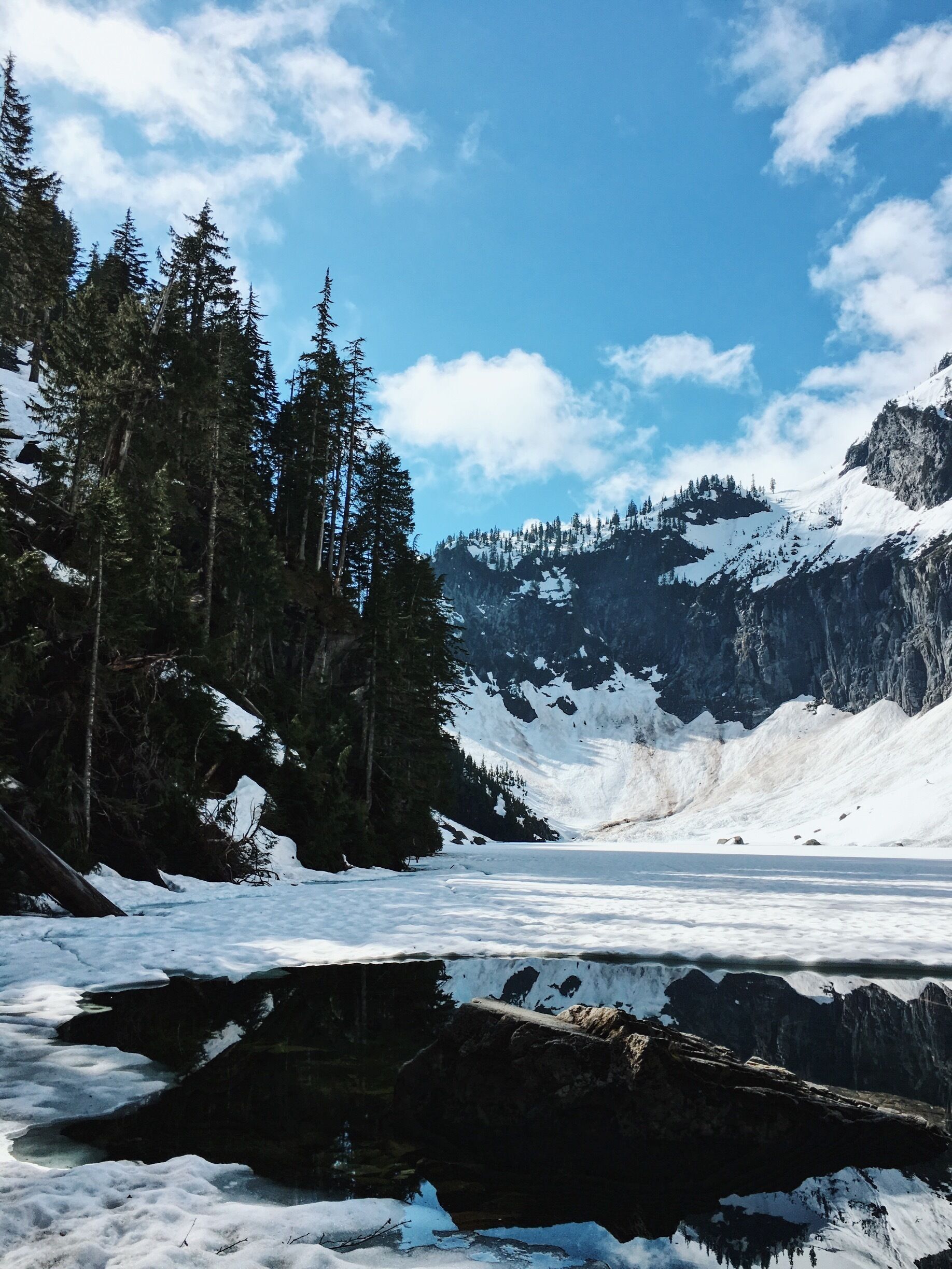 A snow-covered lake surrounded by dense evergreen trees and rugged mountains under a clear blue sky.