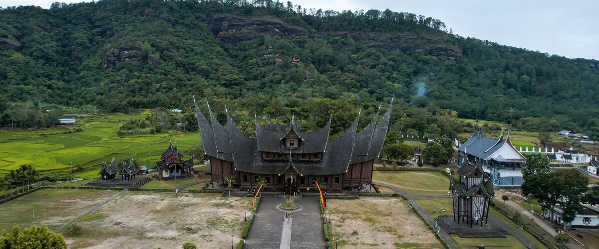A traditional wooden building with a steep, pointed roof, surrounded by terraced rice fields and a forested hill in the background.