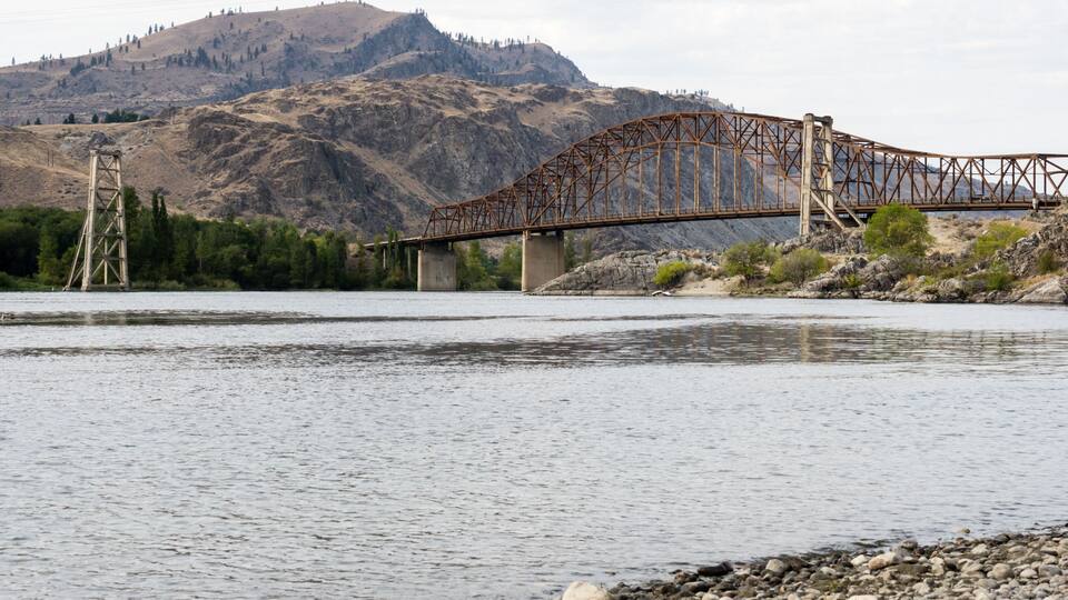A river with a steel bridge, rocky riverbank, and a mountainous backdrop.