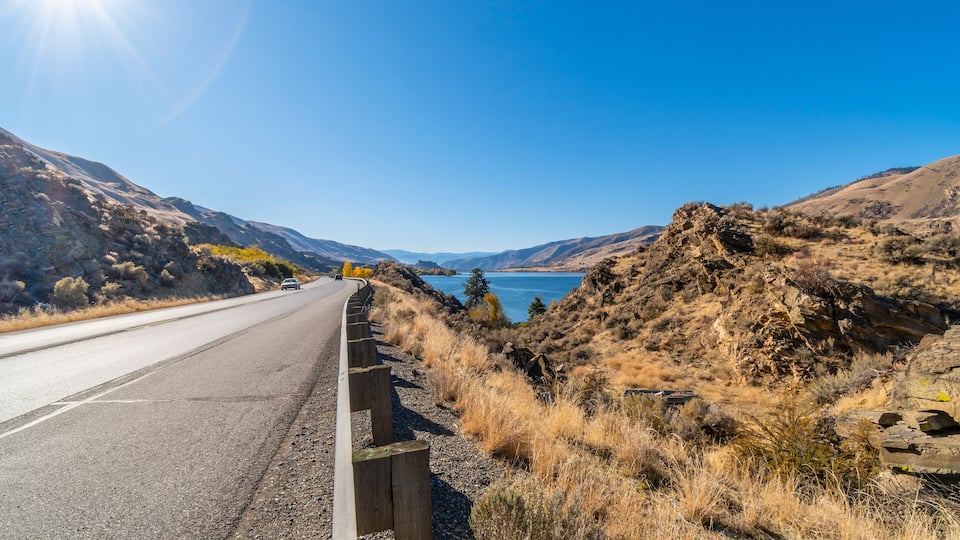 A road with a guardrail, dry grass, rocky hills, and a clear blue sky.