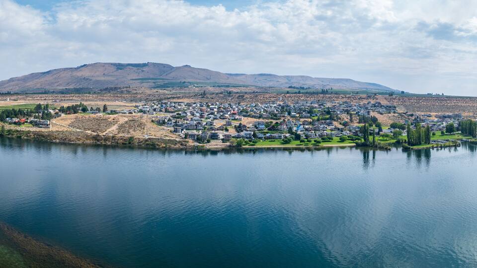A lakeside residential area with houses, a hill in the background, and a clear sky.