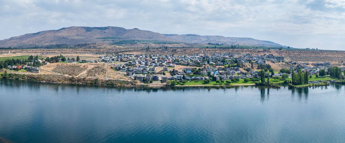 A lakeside residential area with houses, a hill in the background, and a clear sky.
