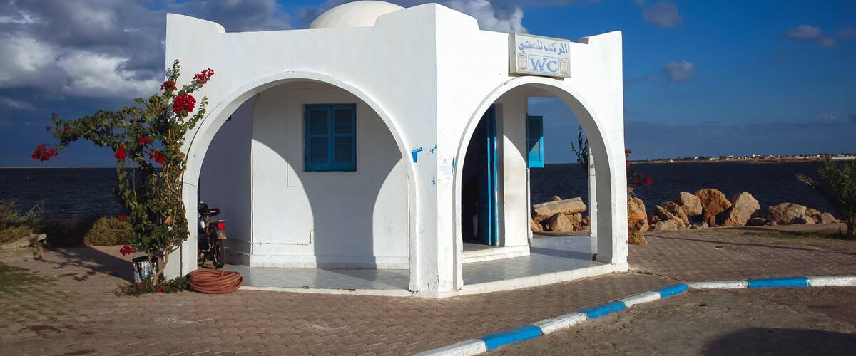 A white building with blue accents, a sign reading "WC," and a view of the sea in the background.