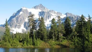 A mountain with snow-capped peaks, a lake reflecting the trees, and a dense forest in the foreground.