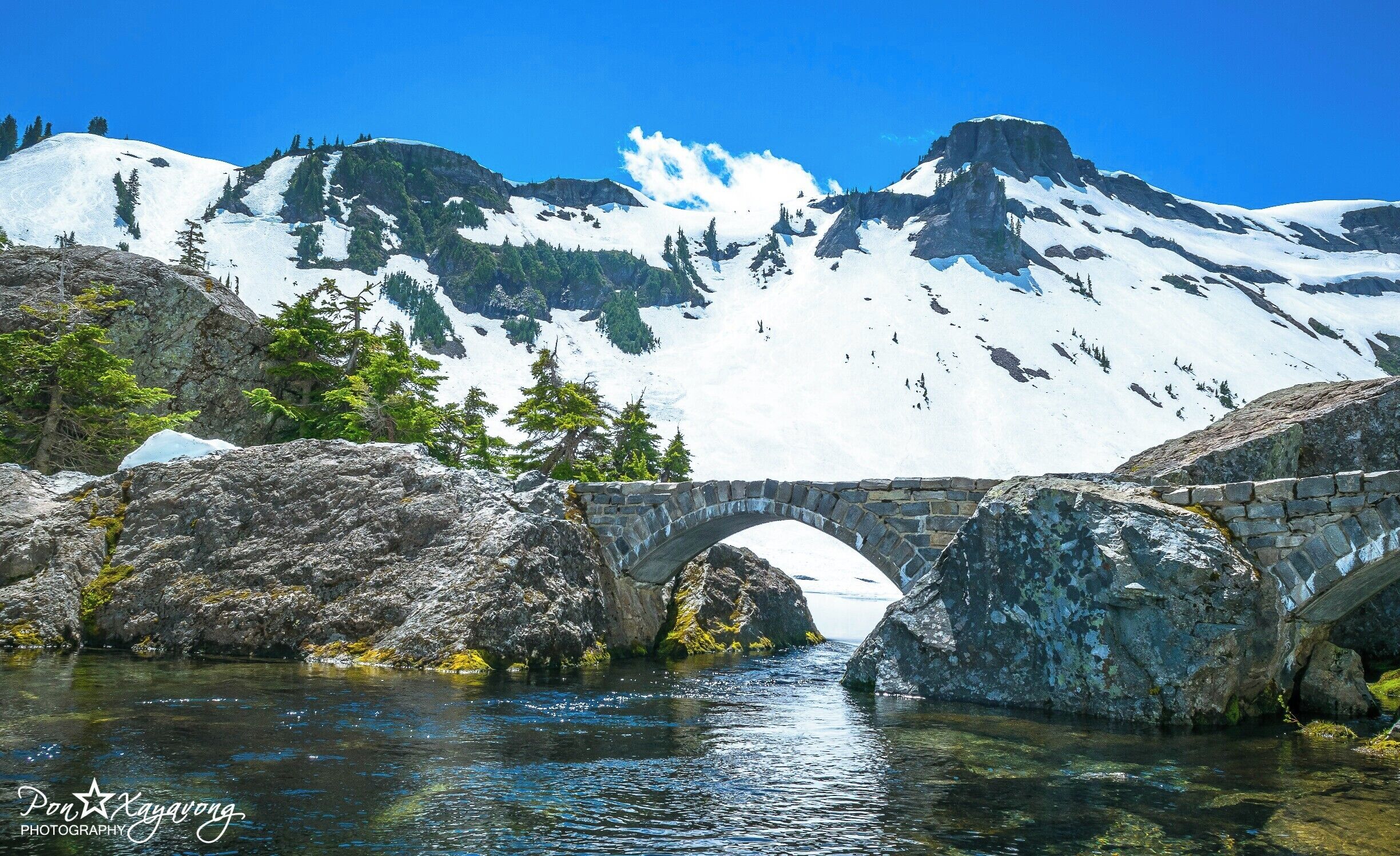 A stone bridge over a clear river, surrounded by snow-capped mountains and green trees.