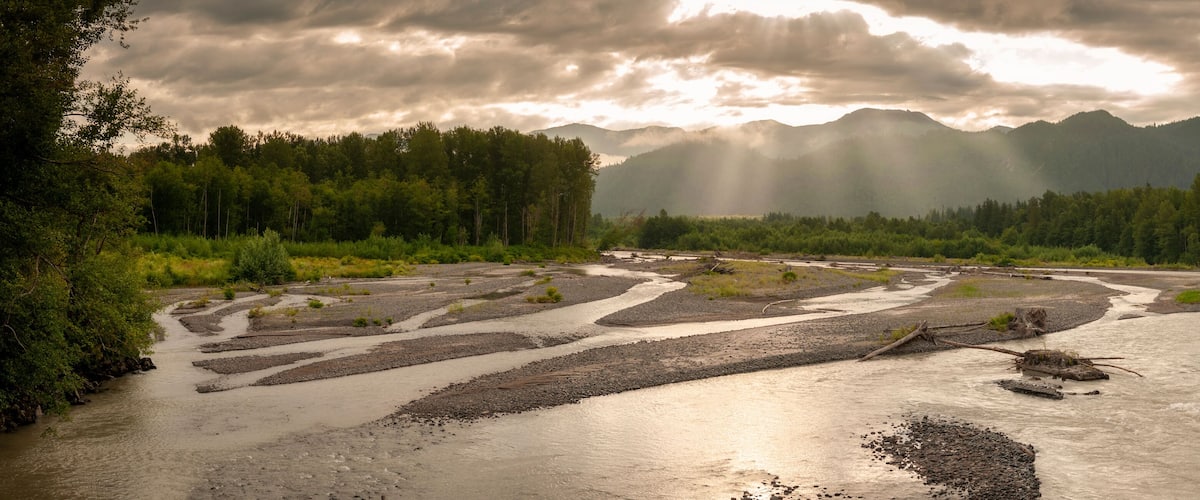 A river winding through a forest with mountains in the background and sunlight filtering through the clouds.