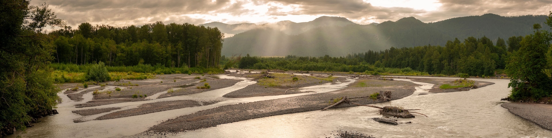 A river winding through a forest with mountains in the background and sunlight filtering through the clouds.