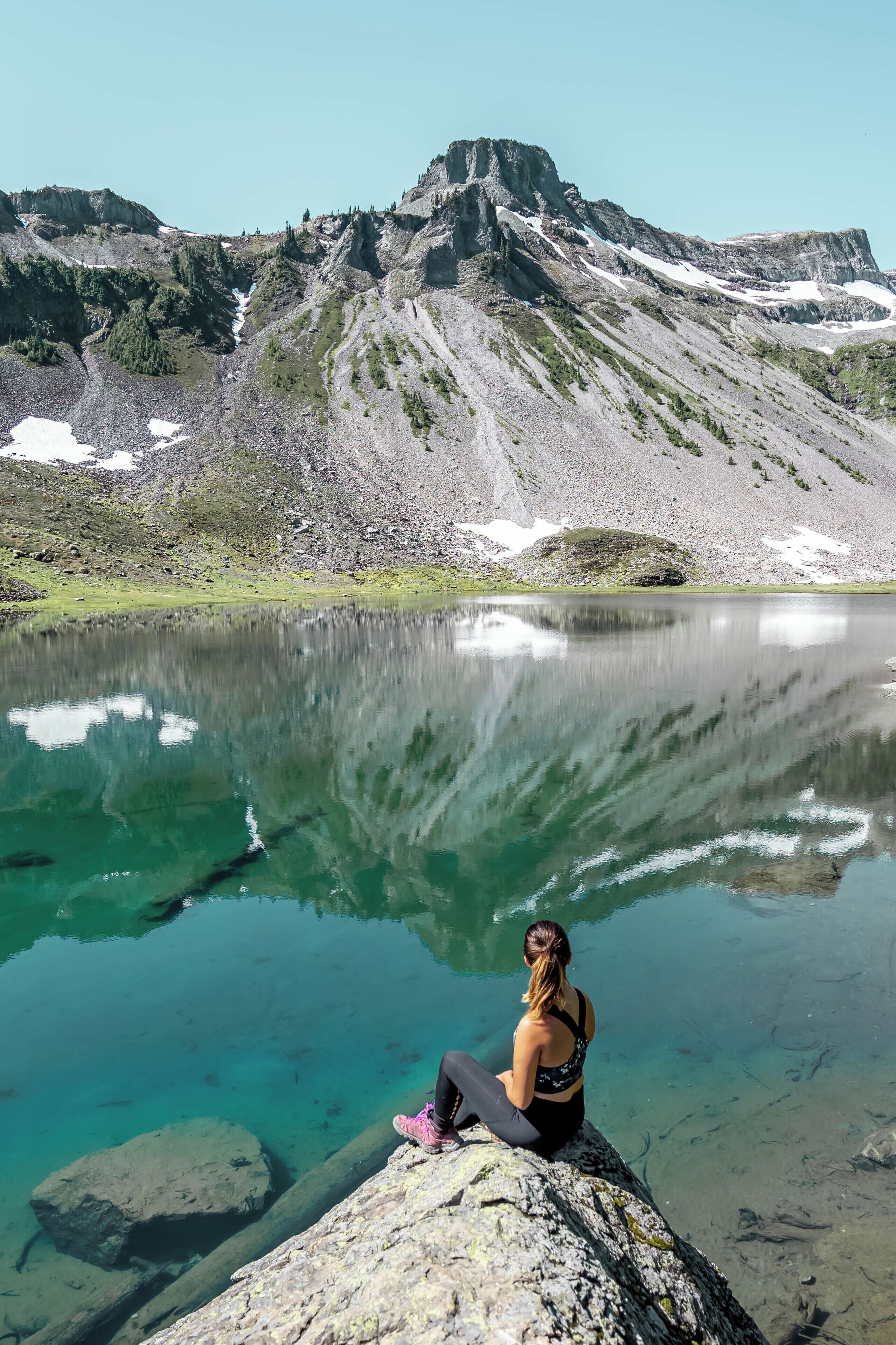 A person sitting on a rock by a clear lake with mountains in the background.