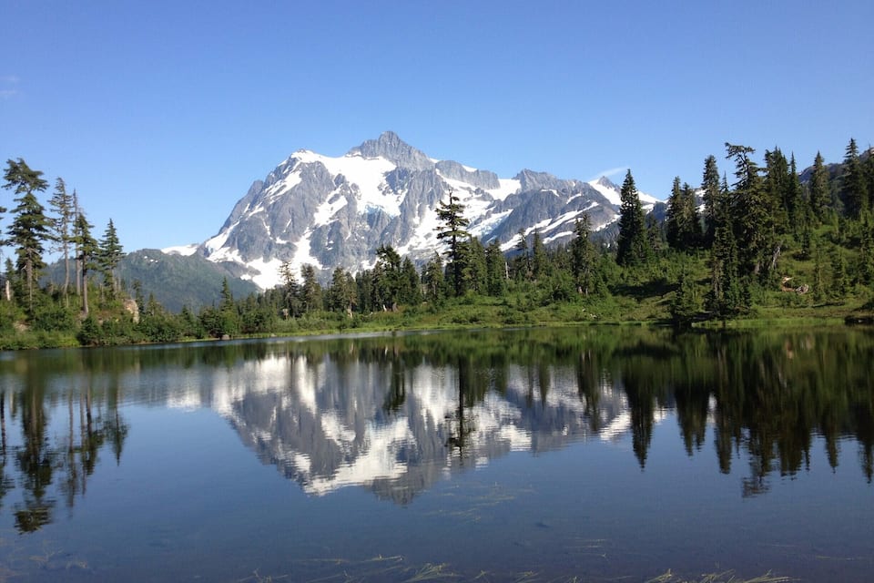 Cordillera con picos nevados, un lago tranquilo que refleja el paisaje y una zona boscosa con árboles de hoja perenne.