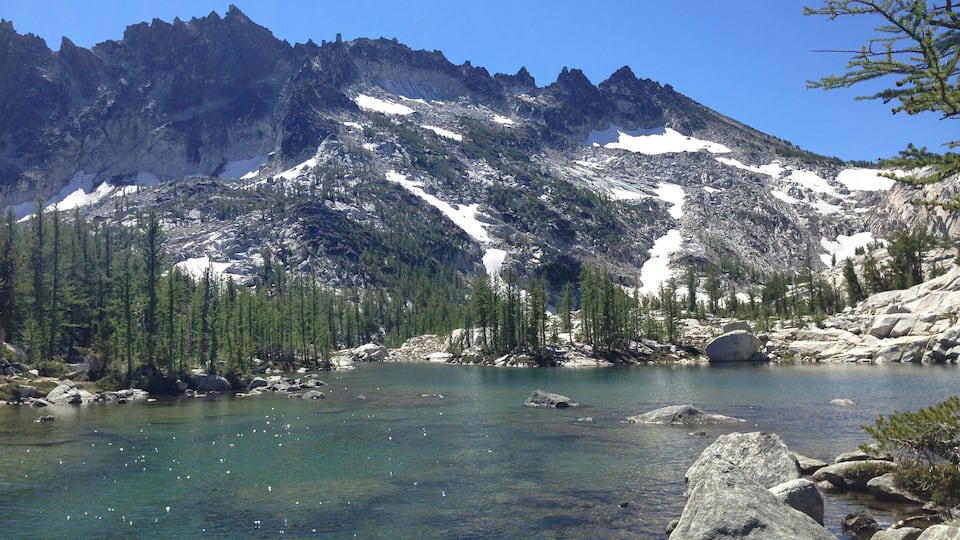 A mountain lake with clear water, surrounded by rocky terrain and snow-capped peaks.
