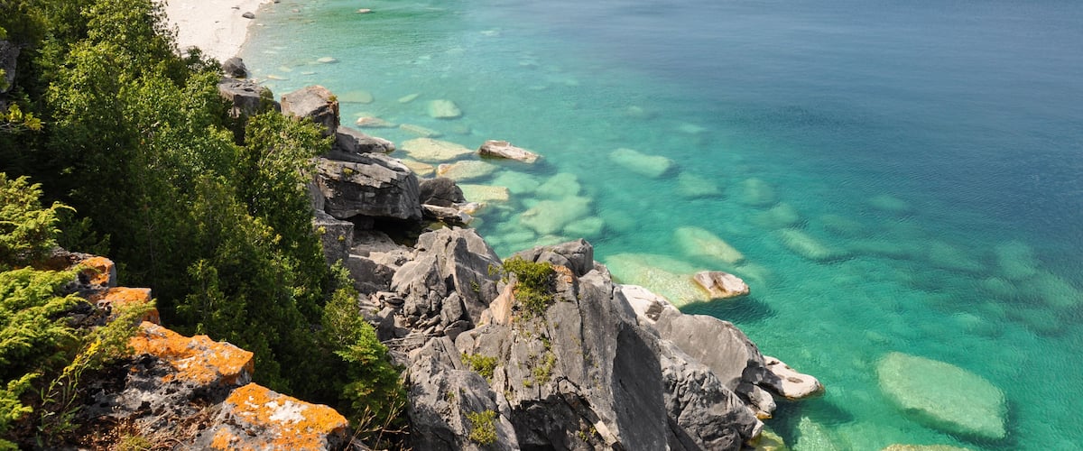 A coastal landscape with a clear turquoise sea, rocky shoreline, and a forested cliff.