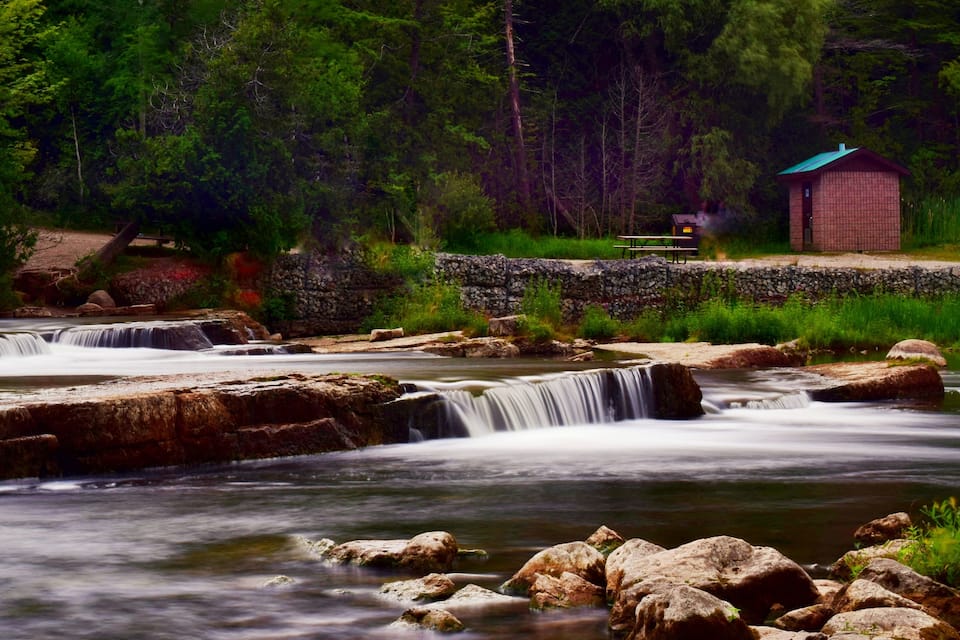 A waterfall, a stone bridge, and a small brick building in a forest.