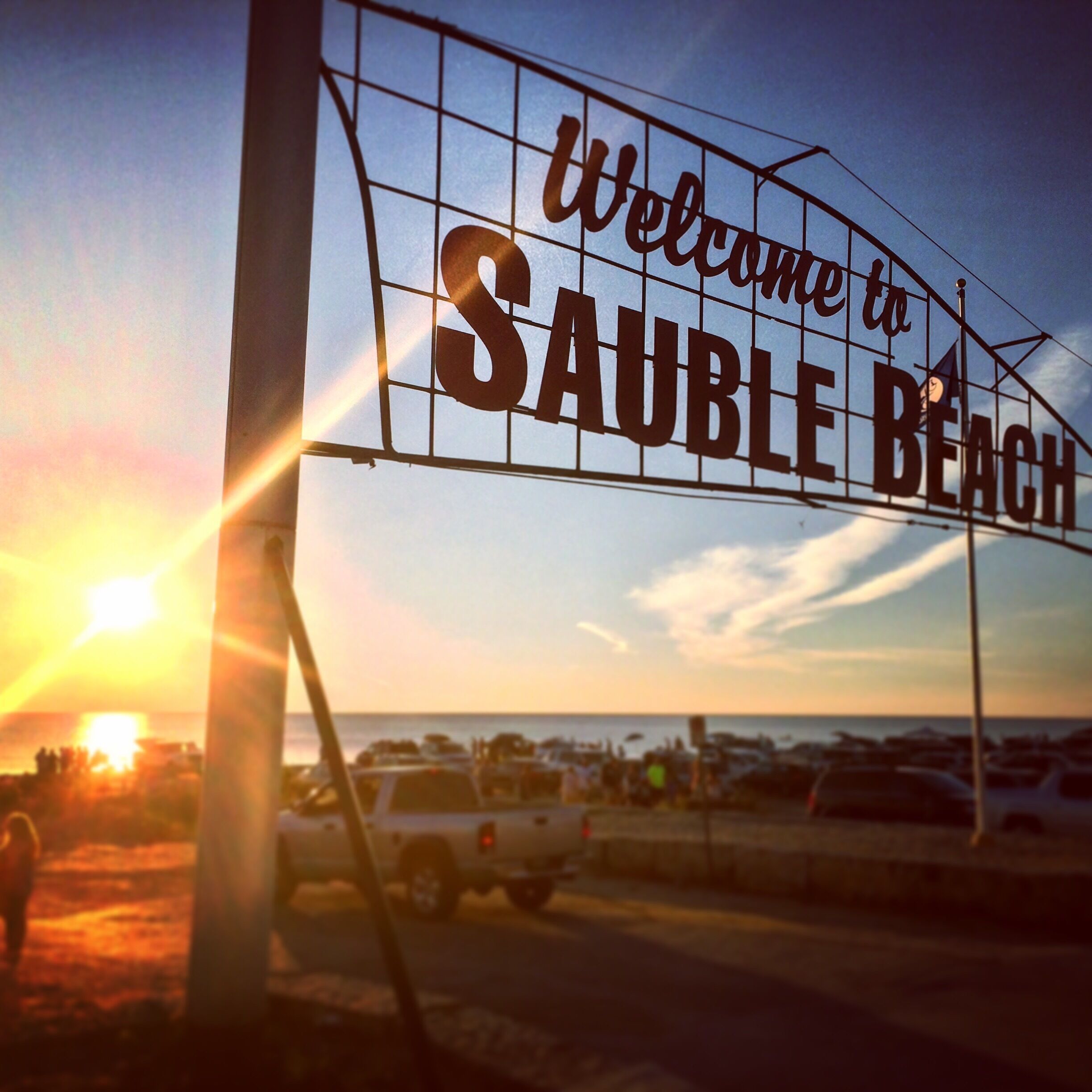 A 'Welcome to Sauble Beach' sign with the sun setting in the background.
