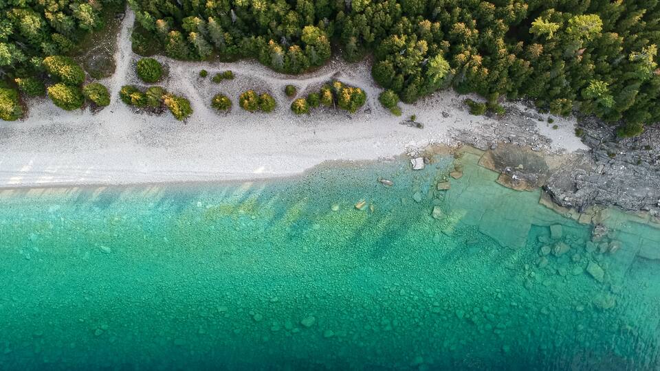 An aerial view of a beach with clear turquoise waters, a sandy shore, and a dense forested area.