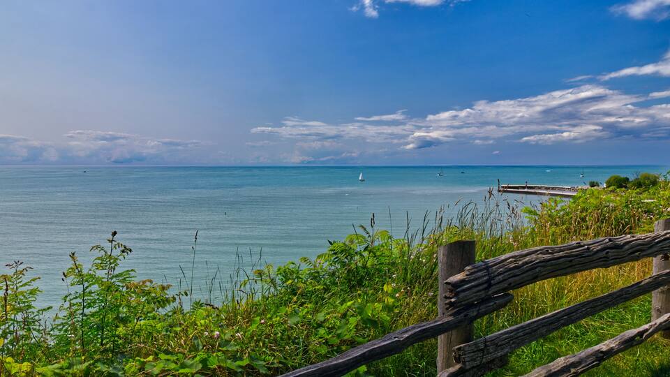 A coastal landscape with a wooden fence, lush greenery, and a calm sea with a sailboat.