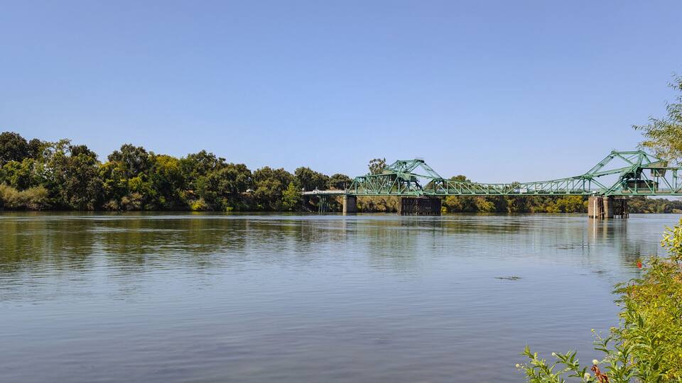 A green metal bridge over a calm river with dense trees on the banks.