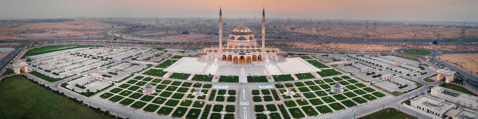 Vista aérea de uma mesquita com um amplo pátio, rodeada por estradas e paisagem desértica.