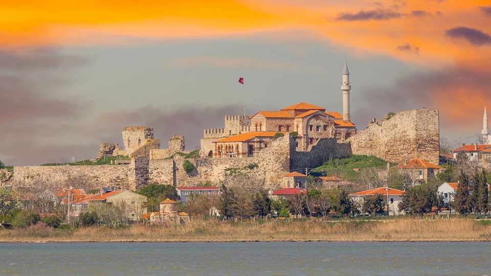 A historic stone fortress with a prominent minaret, surrounded by residential buildings with red roofs, overlooking a body of water.