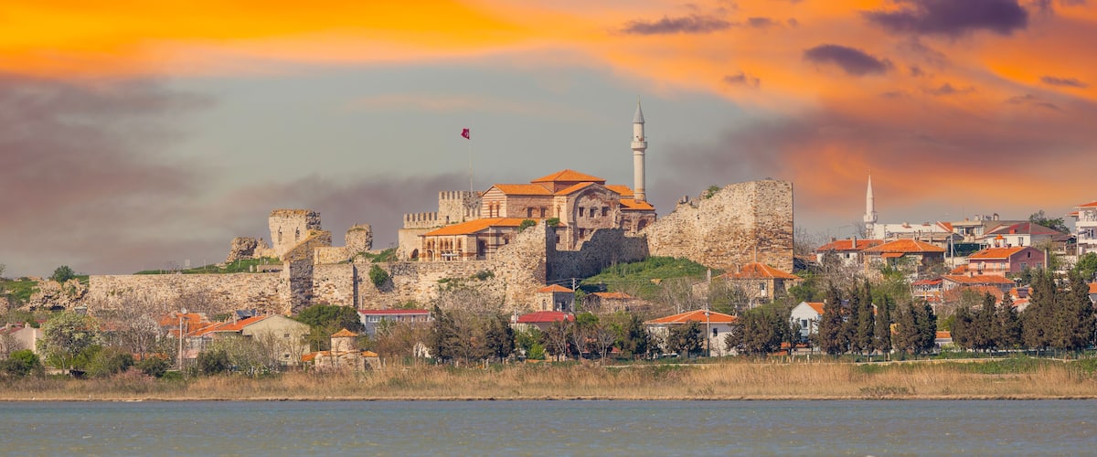 A historic stone fortress with a prominent minaret, surrounded by residential buildings with red roofs, overlooking a body of water.