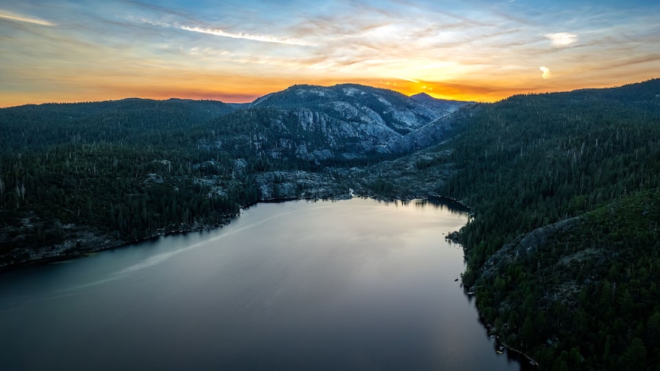 A serene lake surrounded by dense forest and mountains, with a sunset in the background.