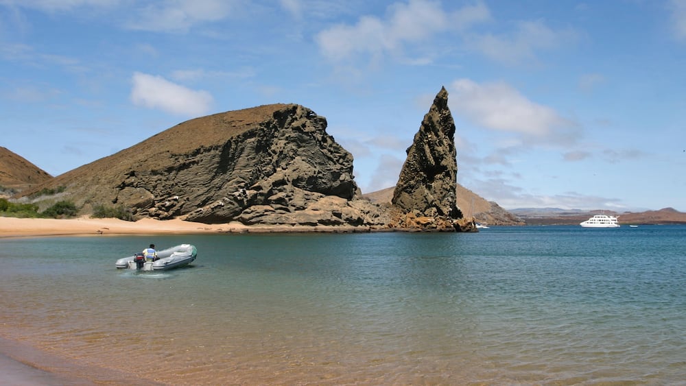 A rocky coastline with a boat in the water and a yacht in the distance.