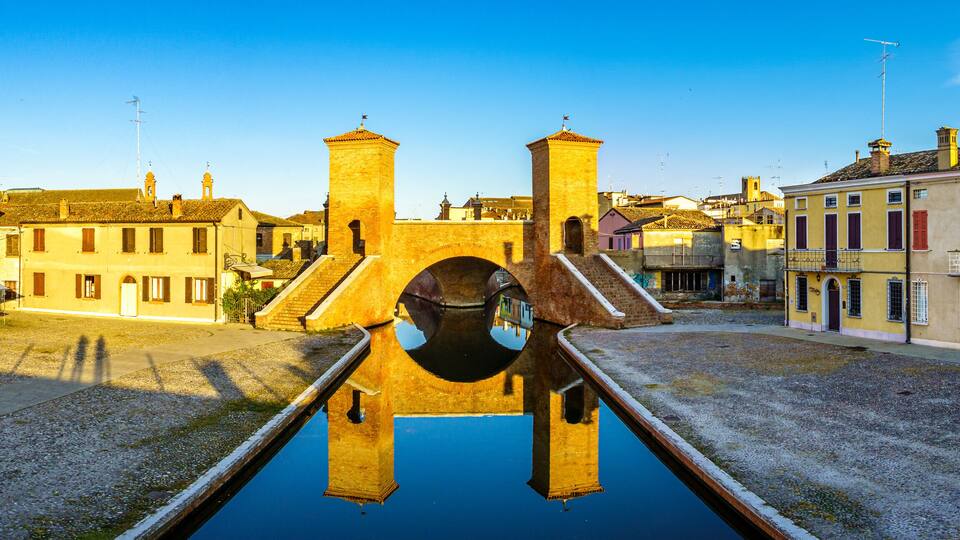 A historic bridge with a reflection in the water, surrounded by colorful buildings.
