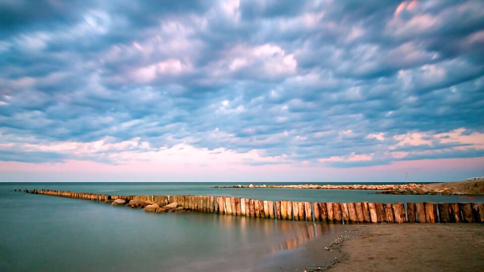 A coastal landscape with a wooden breakwater extending into the sea, under a cloudy sky.