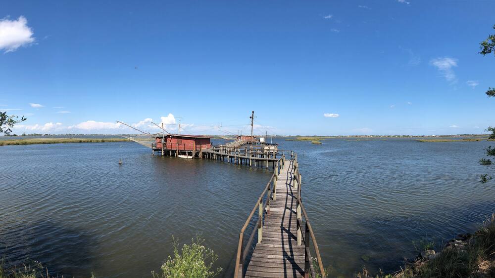 Eine Holzstege führt zu einem roten Gebäude an einem ruhigen See unter wolkenlosem Himmel.
