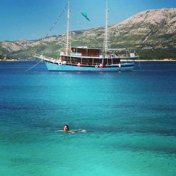 A turquoise boat on calm waters with a swimmer in the foreground and a mountainous landscape in the background.