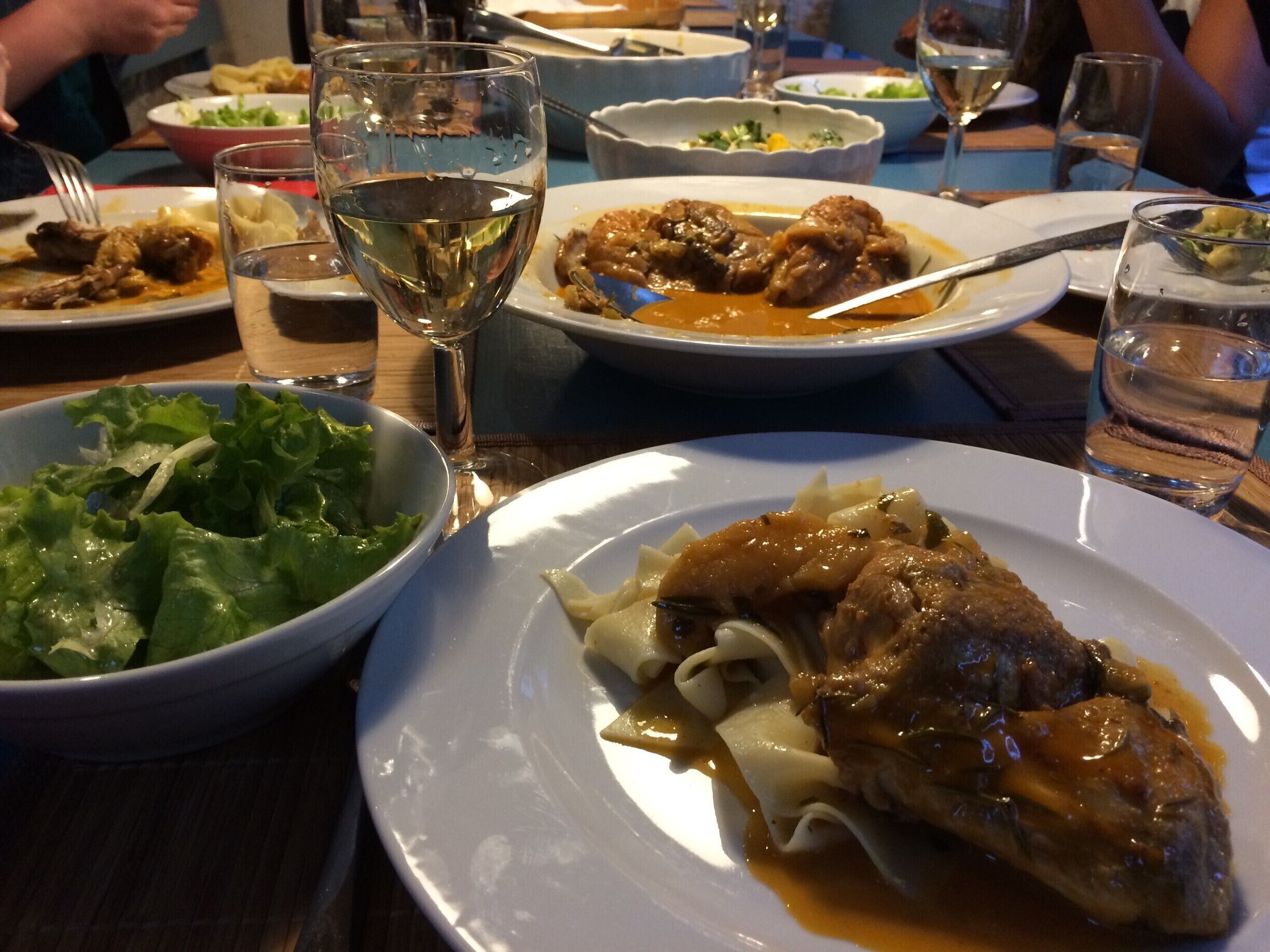 A dining table with various dishes, including a plate of meat with sauce, a bowl of salad, and multiple glasses of water and wine.