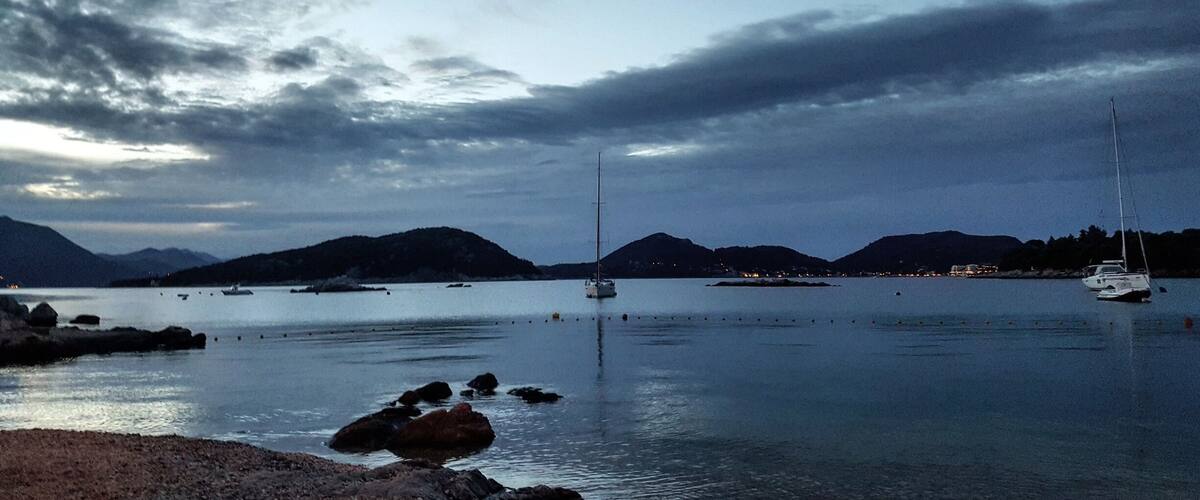 A calm bay with boats, rocky shoreline, and distant mountains under a cloudy sky.