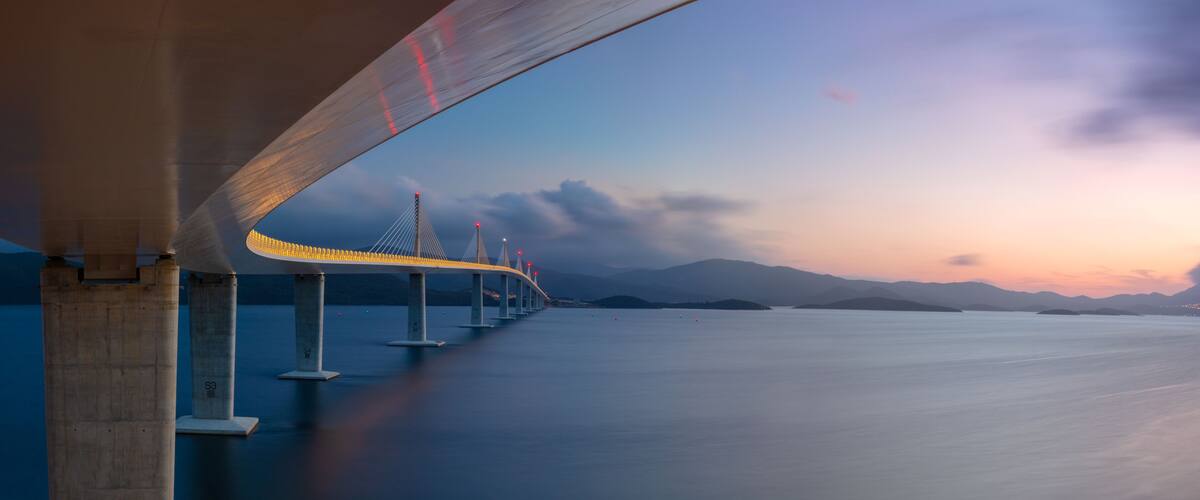 A bridge with illuminated cables, a calm body of water, and distant mountains.