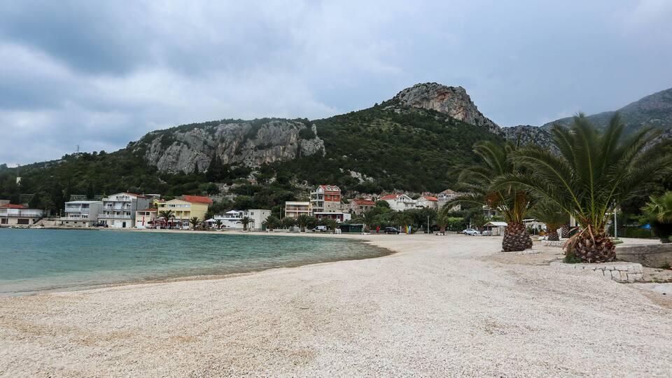 A pebble beach with buildings and a mountain in the background.
