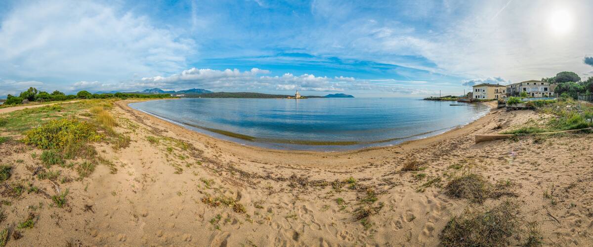 A coastal landscape with a sandy beach, calm sea, and distant mountains under a partly cloudy sky.