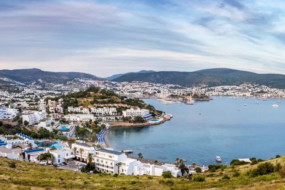 A coastal town with white buildings, a bay with boats, and hills in the background.