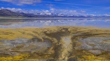 A reflective lake with yellowish water, surrounded by mountains with snow-capped peaks under a clear blue sky.