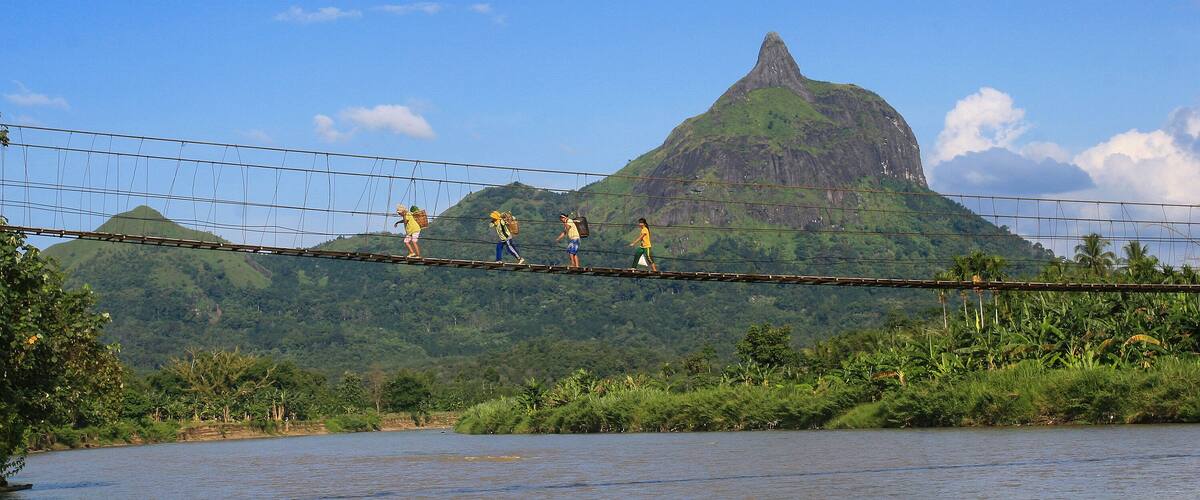 A suspension bridge over a river with people walking across, surrounded by lush greenery and a prominent mountain in the background.